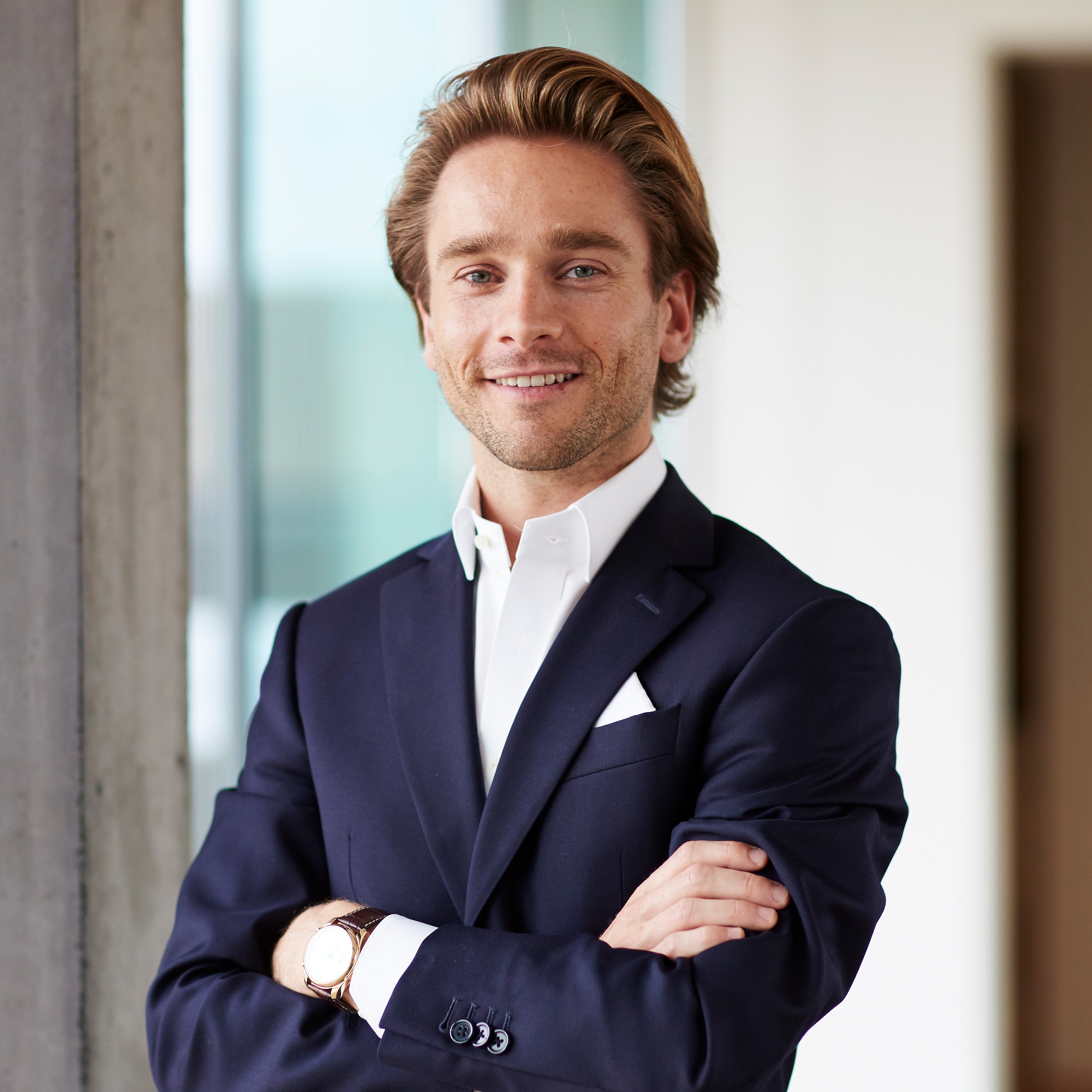 Smiling man in a navy suit, white shirt, and pocket square, arms crossed, with blonde hair.