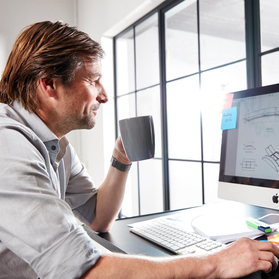 Man in a grey shirt sips from a mug while working on a computer with architectural drawings.